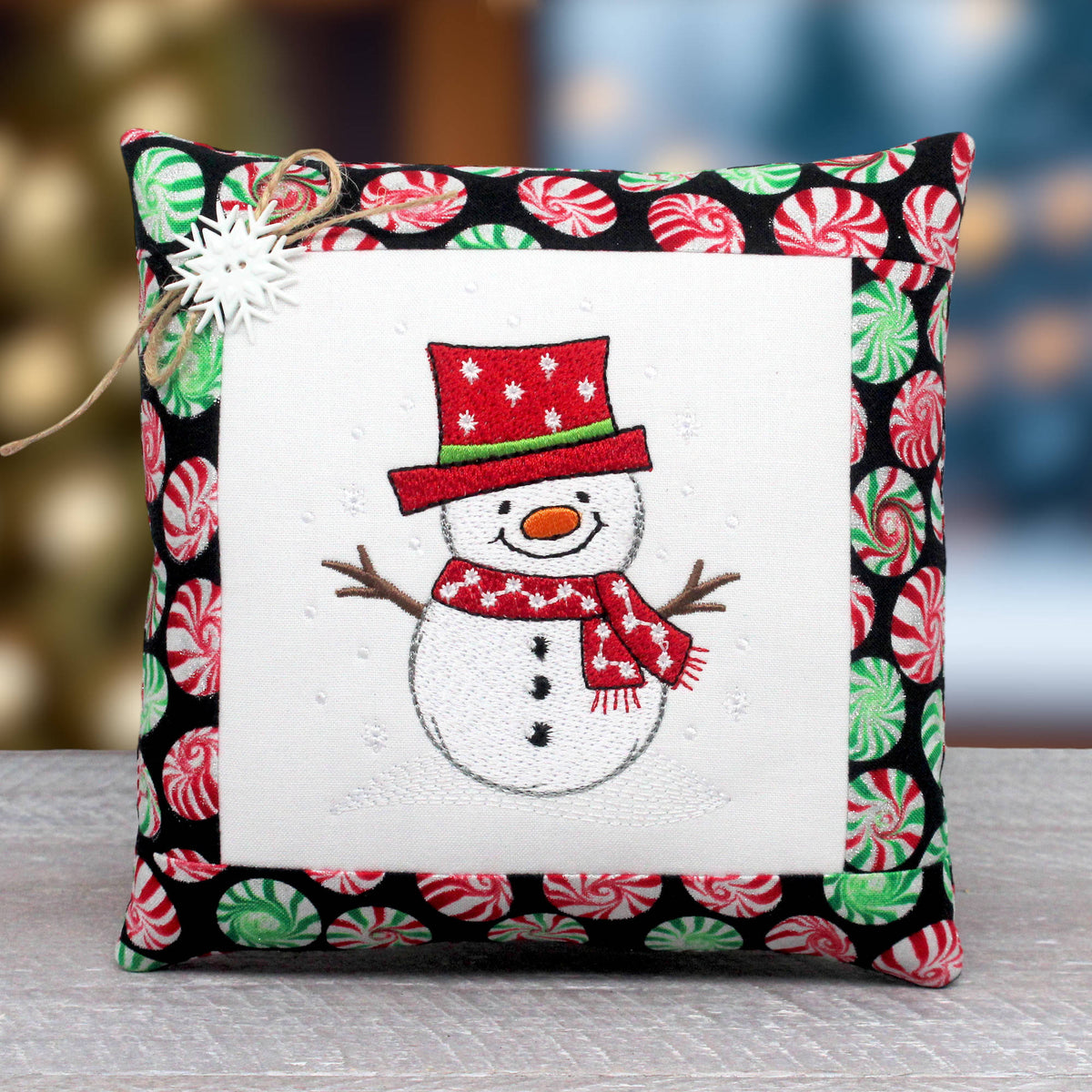 A square decorative pillow with a festive embroidered snowman design in the center, surrounded by a white border and a black border printed with red and green peppermint candies. A small white star ornament and twine are attached to the top left corner.