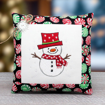 A square decorative pillow with a festive embroidered snowman design in the center, surrounded by a white border and a black border printed with red and green peppermint candies. A small white star ornament and twine are attached to the top left corner.