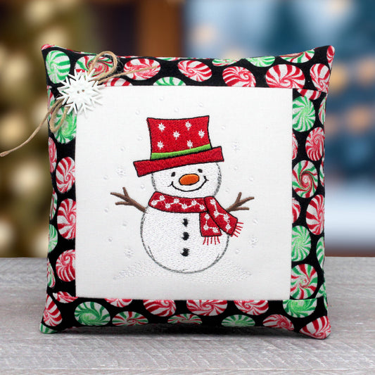A square decorative pillow with a festive embroidered snowman design in the center, surrounded by a white border and a black border printed with red and green peppermint candies. A small white star ornament and twine are attached to the top left corner.