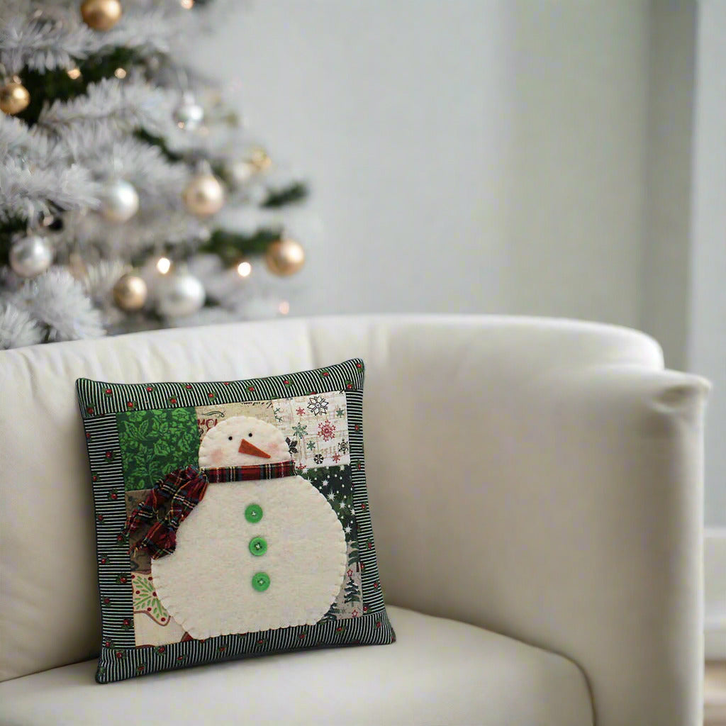 Decorative pillow with a snowman design on a white couch with a Christmas tree in the background.