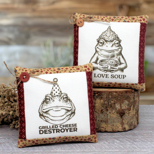 Two decorative square fabric mini pillows displayed on a rustic wood stump. The front pillow reads 'Grilled Cheese Destroyer' with a frog illustration and a party hat, and the back pillow reads 'I Love Soup' with a frog holding a bowl.