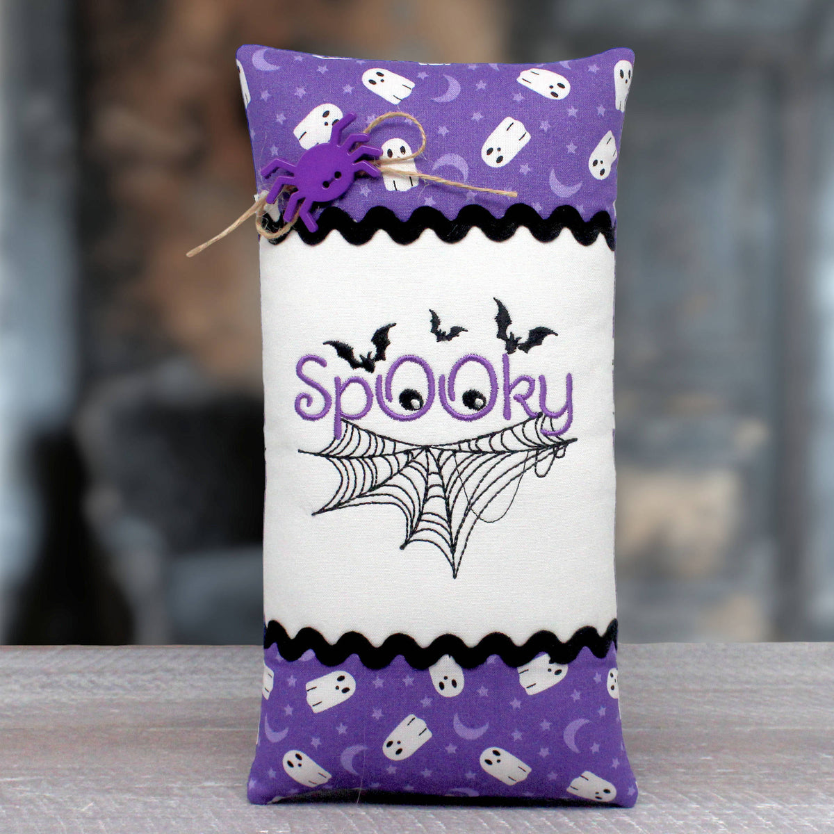 A rectangular fabric spooktacular pillow with a purple top and bottom patterned border featuring ghosts, moons, and bats, a white center, black scalloped trim, embroidered black spiderwebs and bats, purple 'Spooky' lettering, and a purple spider decoration tied with twine.