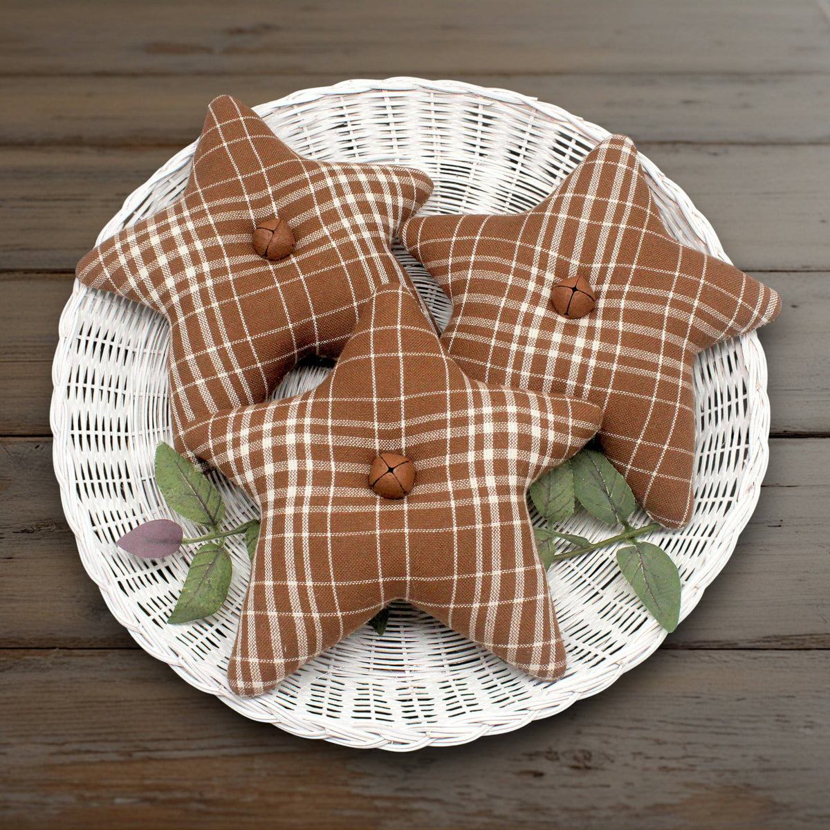 Three star homespun bowl fillers in a brown and white plaid pattern, arranged on a white wicker tray with green leaves on a rustic wooden table.