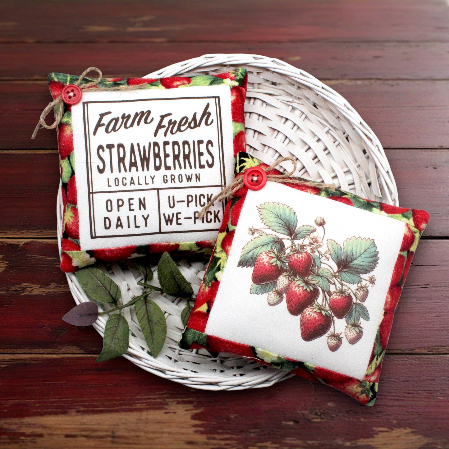Two decorative fabric strawberry-themed mini pillows displayed on a white woven tray over a dark wooden surface. One pillow shows a vintage-style label reading 'Farm Fresh Strawberries Locally Grown' with red strawberries and green leaves around the border.