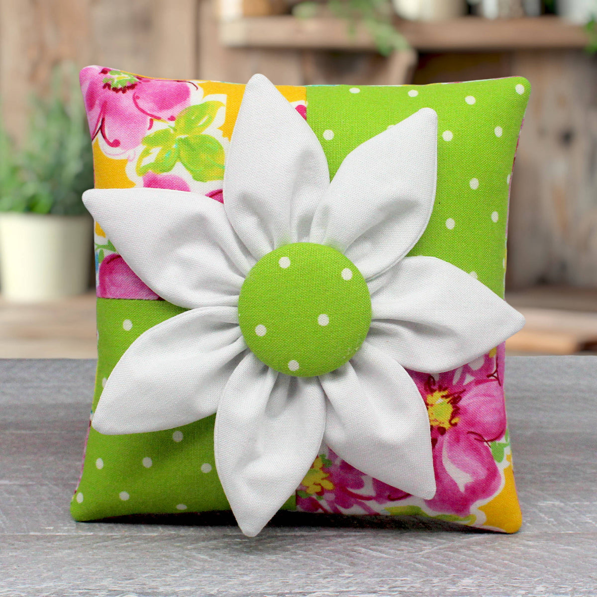 A small square decorative pillow with a colorful floral and polka-dot fabric background, featuring a large white fabric flower applique with a green polka-dot center on the front.