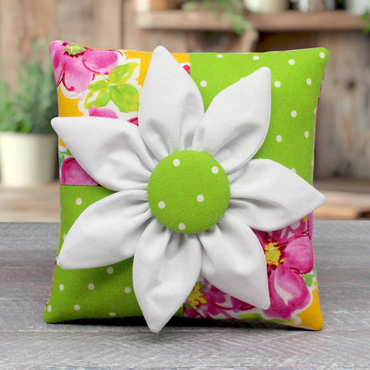 A small square decorative pillow with a colorful floral and polka-dot fabric background, featuring a large white fabric flower applique with a green polka-dot center on the front.