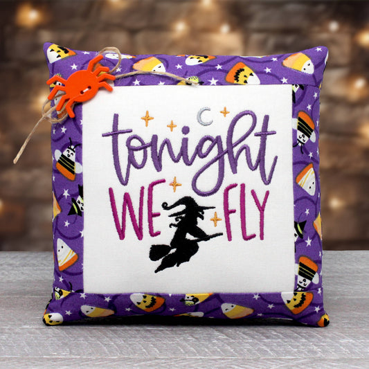 Mini pillow tuck or bowl filler is embroidered with the words 'tonight we fly' alongside a witch on a broom.  The center panel is trimmed with a Halloween print fabric with candy corn on a purple background.