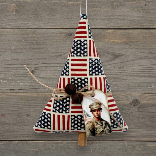 A fabric hanging ornament decorated with an American flag patchwork pattern, twine, two rusty bells, and a printed tag of a soldier, shown against a rustic gray wooden plank background.