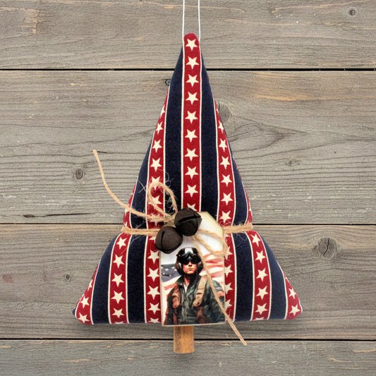 A patriotic Christmas ornament shaped like a Christmas tree, made from red, white, and navy blue striped fabric with white stars, decorated with three small rusty bells, twine, and a printed photo of a American service member, hanging against a rustic gray wooden plank background.