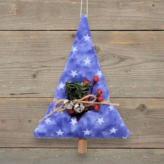 A handmade fabric Christmas tree ornament in a blue watercolor pattern with white stars, decorated with a small pine sprig, red berries, and silver bells, hanging against a rustic wooden plank background.
