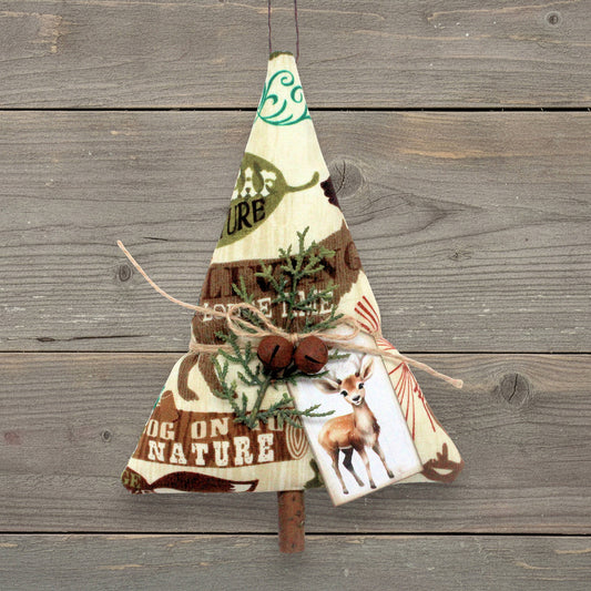 A rustic Christmas tree ornament made from fabric, featuring brown and cream tones, pine branches, twine, and a deer print, hanging against a weathered gray wood plank background.