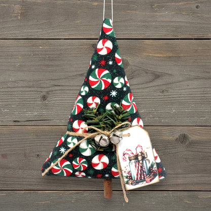 A handmade Christmas tree ornament made from dark fabric with a candy cane and snowflake pattern, decorated with small green pine branches, twine, silver bells, and a hanging gift tag, shown against a rustic wooden plank background.