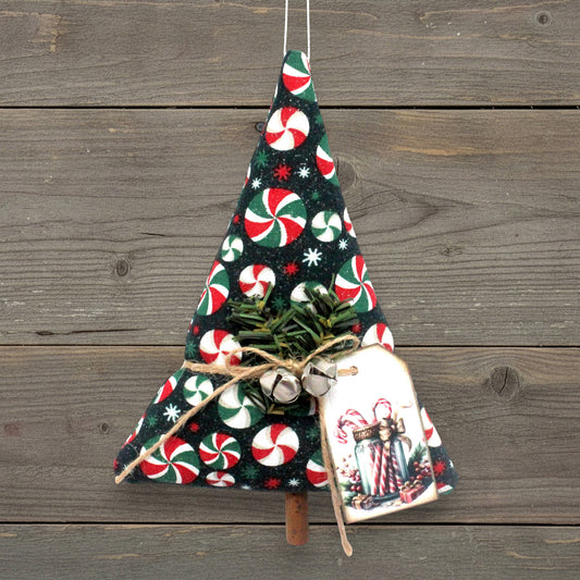 A handmade Christmas tree ornament made from dark fabric with a candy cane and snowflake pattern, decorated with small green pine branches, twine, silver bells, and a hanging gift tag, shown against a rustic wooden plank background.