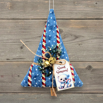 A handmade Christmas tree-shaped hanging ornament made from blue fabric with white polka dots, red and white candy cane stripes, gold bells, twine, and a 'Sweet Seasons!' gift tag, displayed against a rustic wooden plank background.
