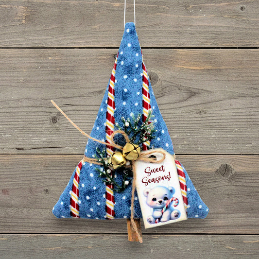 A handmade Christmas tree-shaped hanging ornament made from blue fabric with white polka dots, red and white candy cane stripes, gold bells, twine, and a 'Sweet Seasons!' gift tag, displayed against a rustic wooden plank background.