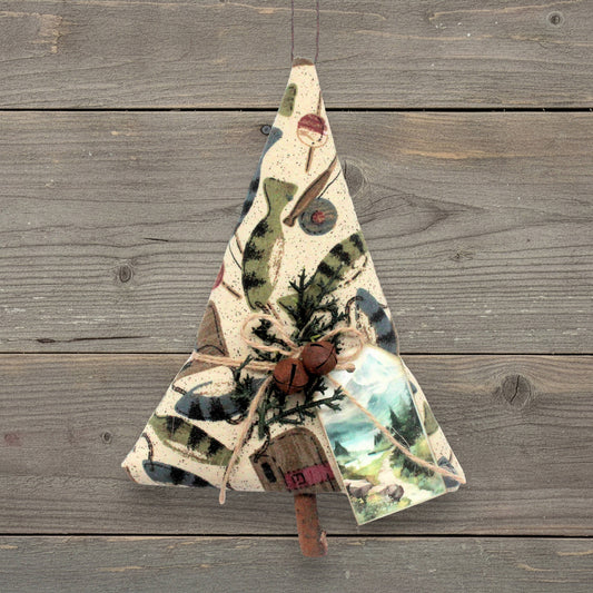 A handmade fabric Christmas tree ornament hanging against a rustic gray wooden plank background. The ornament is made from cream-colored fabric printed with green pine trees, rusty bells, and a small vintage-style tag, suspended by a string.