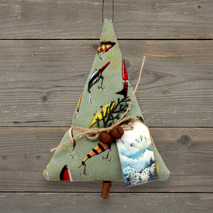 A handmade ornament or made from sage green fabric, decorated with fishing lures, hooks, twigs, rusty bells, and a small printed tag showing a mountain scene. It hangs against a rustic gray wooden plank background