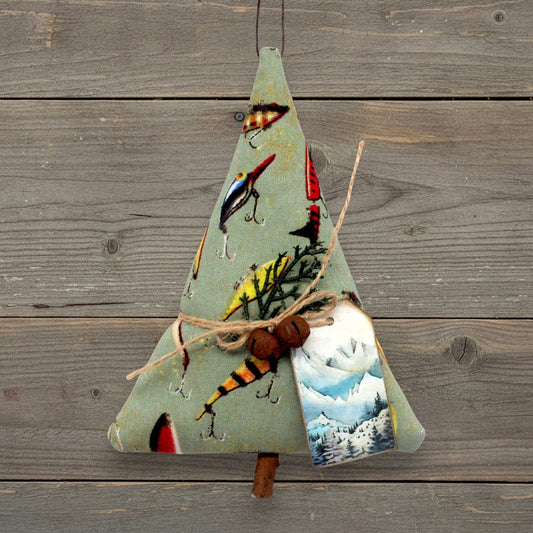 A handmade ornament or made from sage green fabric, decorated with fishing lures, hooks, twigs, rusty bells, and a small printed tag showing a mountain scene. It hangs against a rustic gray wooden plank background