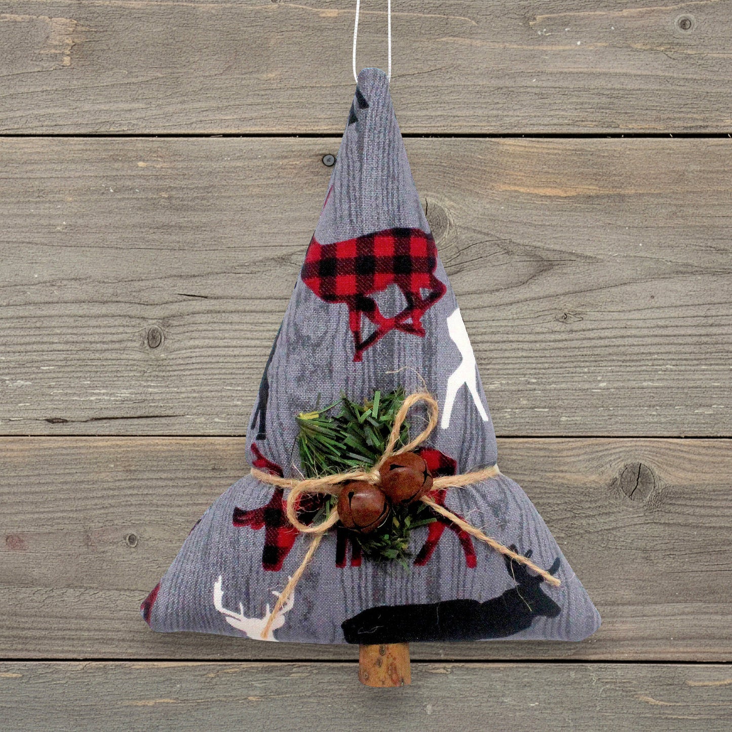 A fabric Christmas tree ornament made from gray corduroy with red and black buffalo plaid forest animals, green pine branches, and two rusty bells tied with twine, hanging against a rustic wood plank background.