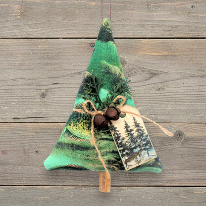 A fabric Christmas tree ornament with a green forest landscape print tag, decorated with pine branches, two rusty bells, a twine bow, and a small printed tag, hanging against a rustic wood plank background.