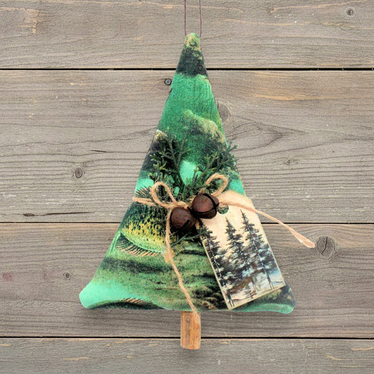 A fabric Christmas tree ornament with a green forest landscape print tag, decorated with pine branches, two rusty bells, a twine bow, and a small printed tag, hanging against a rustic wood plank background.