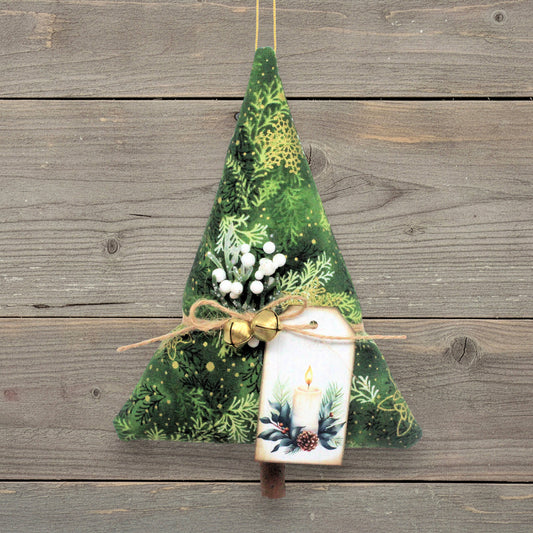 A Christmas tree-shaped fabric ornament in green botanical print, hanging against a rustic gray wooden plank background. It has a tag with a printed candle illustration, tied with twine and gold bells.