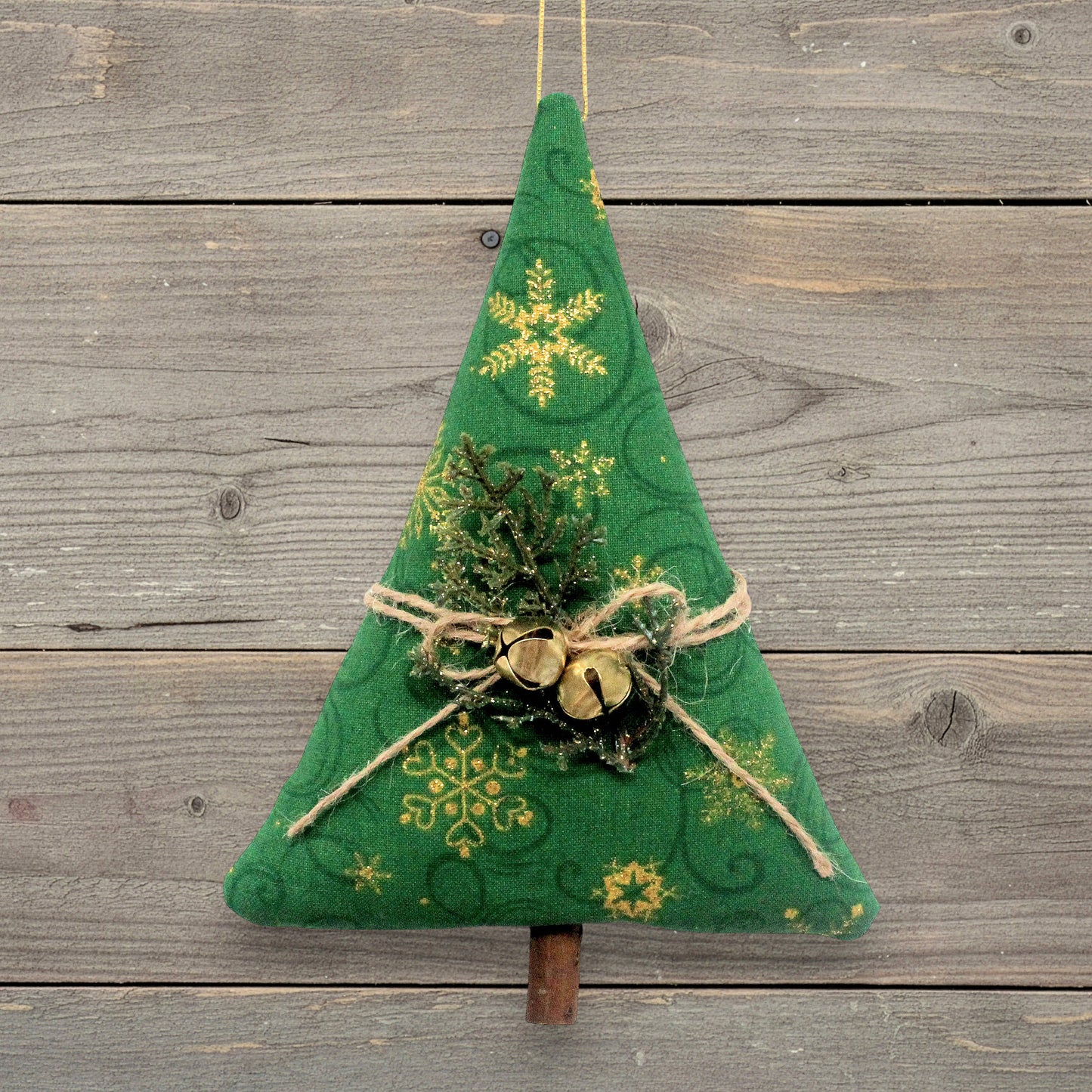 A handmade Christmas tree ornament with gold snowflake patterns, twine bow, small gold bells, and faux greenery, hanging against a rustic gray wooden plank background.