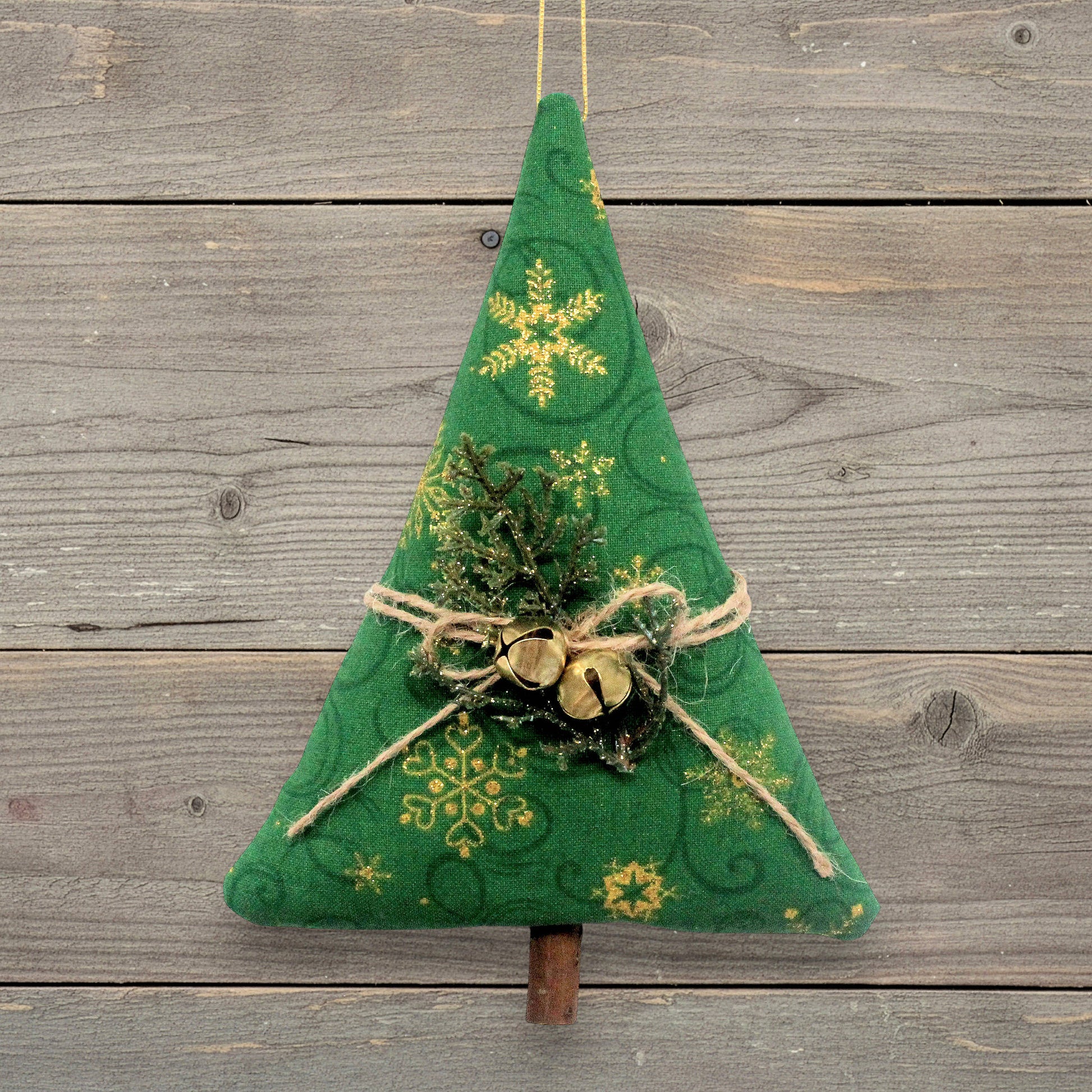A handmade Christmas tree ornament with gold snowflake patterns, twine bow, small gold bells, and faux greenery, hanging against a rustic gray wooden plank background.