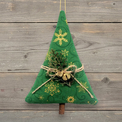 A handmade Christmas tree ornament with gold snowflake patterns, twine bow, small gold bells, and faux greenery, hanging against a rustic gray wooden plank background.