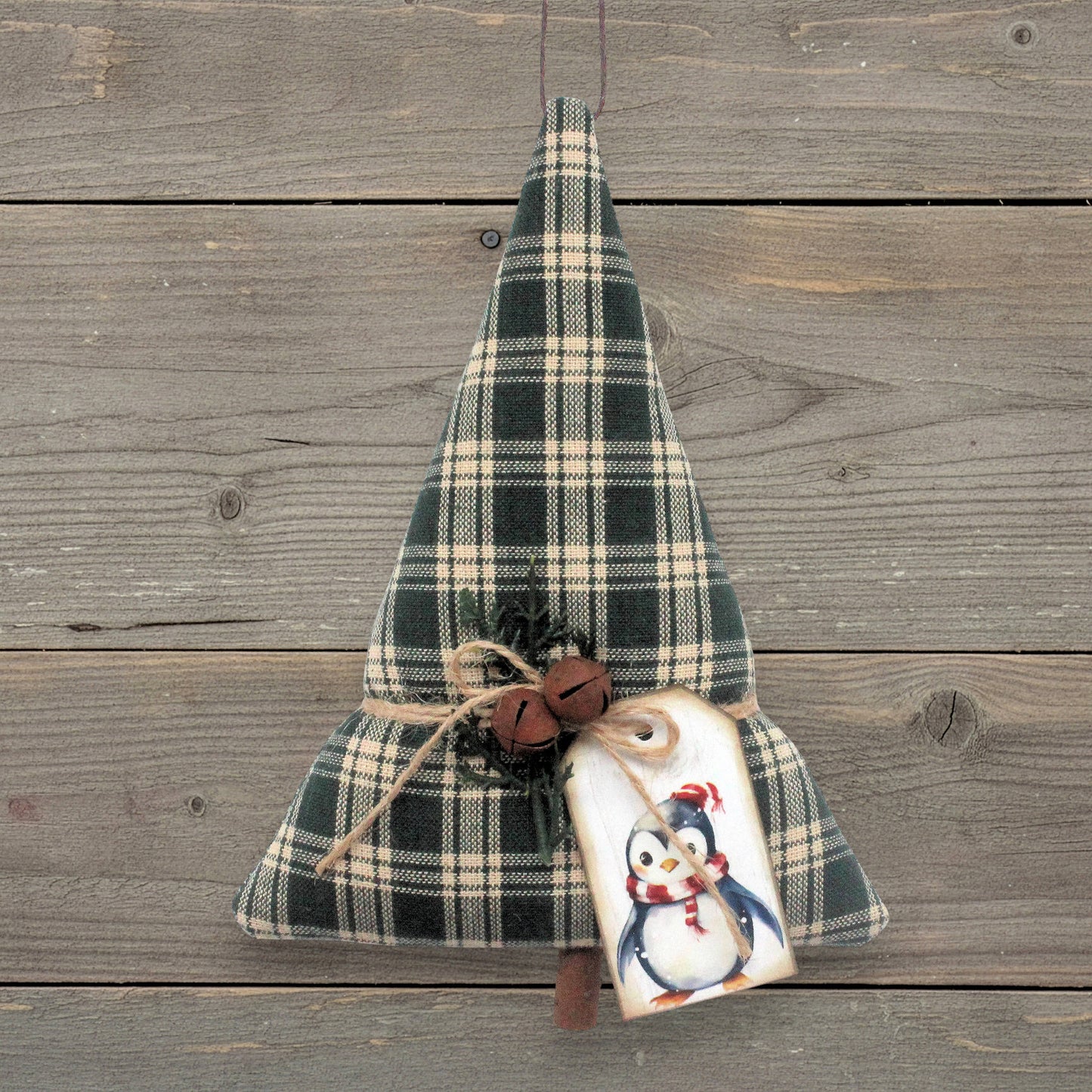 A handmade Christmas tree ornament made from dark green and beige plaid fabric, tied with twine and decorated with a penguin tag and small bells, hanging against a rustic wooden plank background.