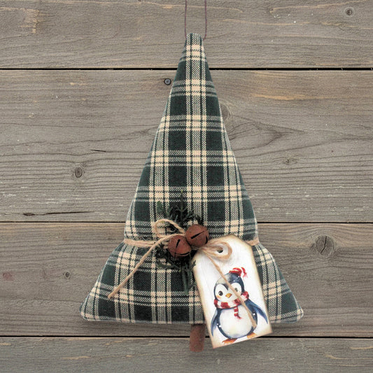 A handmade Christmas tree ornament made from dark green and beige plaid fabric, tied with twine and decorated with a penguin tag and small bells, hanging against a rustic wooden plank background.