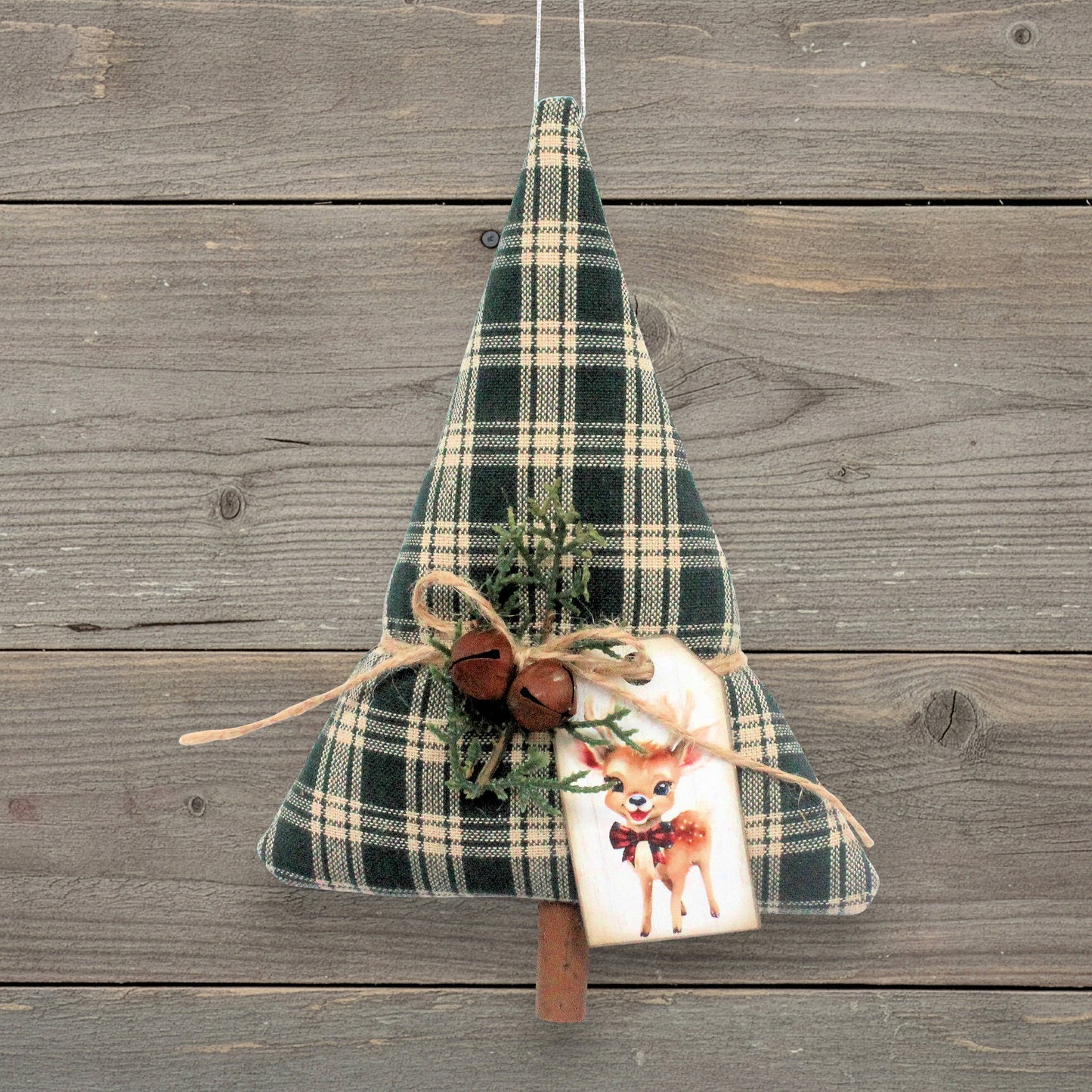 A Christmas tree-shaped hanging decoration made of dark green and beige plaid fabric, decorated with twine, small rusty bells, evergreen sprigs, and a white gift tag featuring a deer illustration, hanging against a rustic gray wooden plank background.