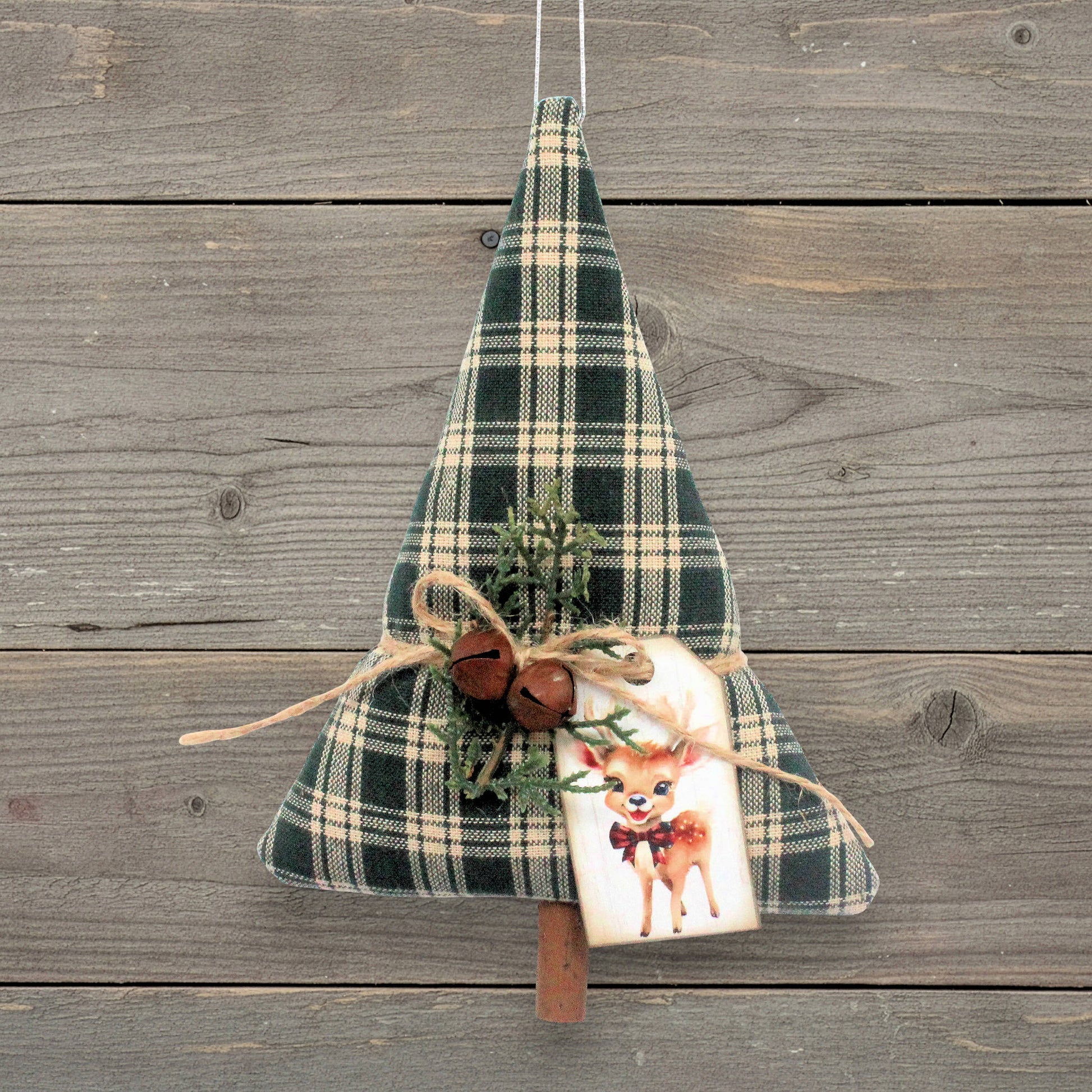 A Christmas tree-shaped hanging decoration made of dark green and beige plaid fabric, decorated with twine, small rusty bells, evergreen sprigs, and a white gift tag featuring a deer illustration, hanging against a rustic gray wooden plank background.