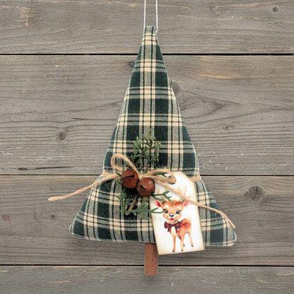 A Christmas tree-shaped hanging decoration made of dark green and beige plaid fabric, decorated with twine, small rusty bells, evergreen sprigs, and a white gift tag featuring a deer illustration, hanging against a rustic gray wooden plank background.