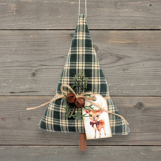 A Christmas tree-shaped hanging decoration made of dark green and beige plaid fabric, decorated with twine, small rusty bells, evergreen sprigs, and a white gift tag featuring a deer illustration, hanging against a rustic gray wooden plank background.
