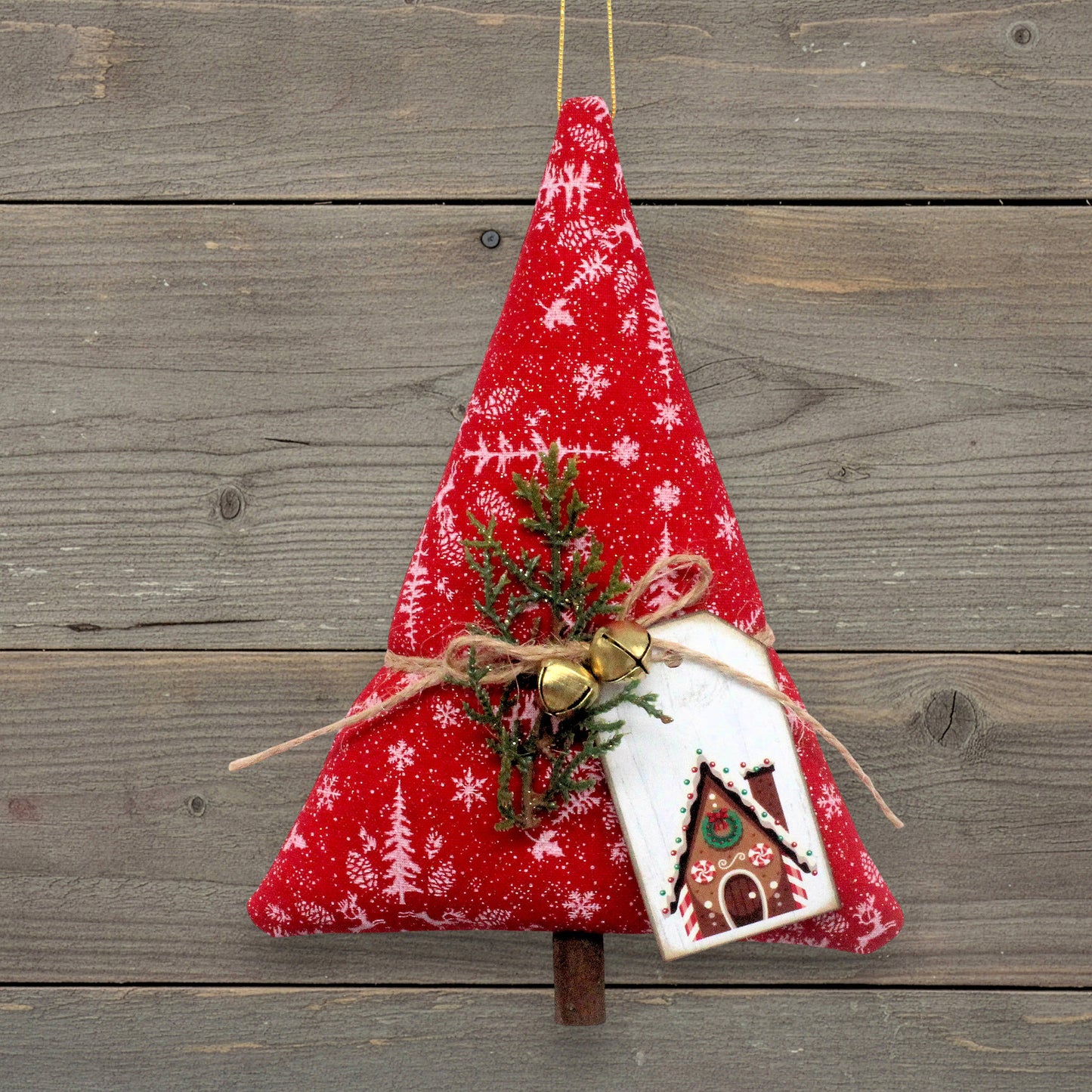 A red fabric Christmas tree ornament with white snowflake and tree patterns, decorated with a small pine sprig, gold jingle bells, twine, and a white gingerbread house tag, hanging against a rustic gray wooden plank background.