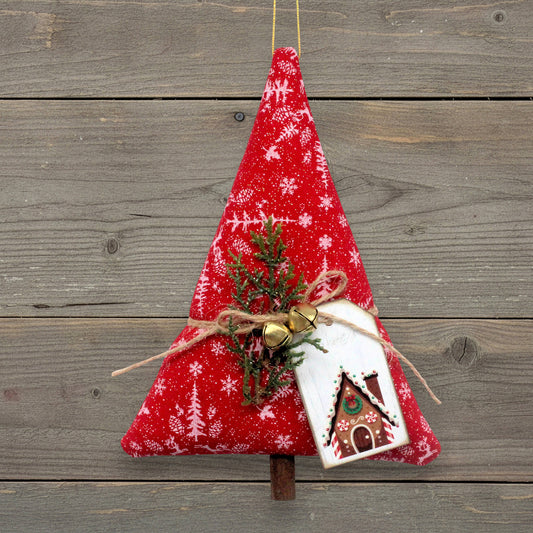 A red fabric Christmas tree ornament with white snowflake and tree patterns, decorated with a small pine sprig, gold jingle bells, twine, and a white gingerbread house tag, hanging against a rustic gray wooden plank background.