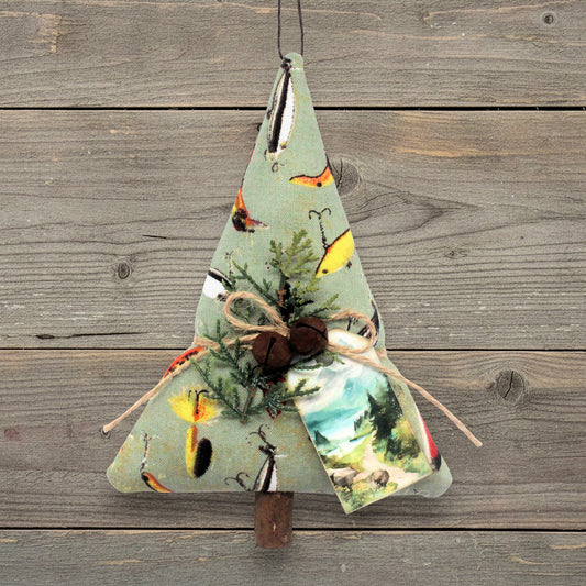 A handmade fabric Christmas tree ornament with a light sage green background, printed with rustic fly fishing and landscape motifs, decorated with rusty bells, greenery, and a bow, hanging against a weathered wood plank background.
