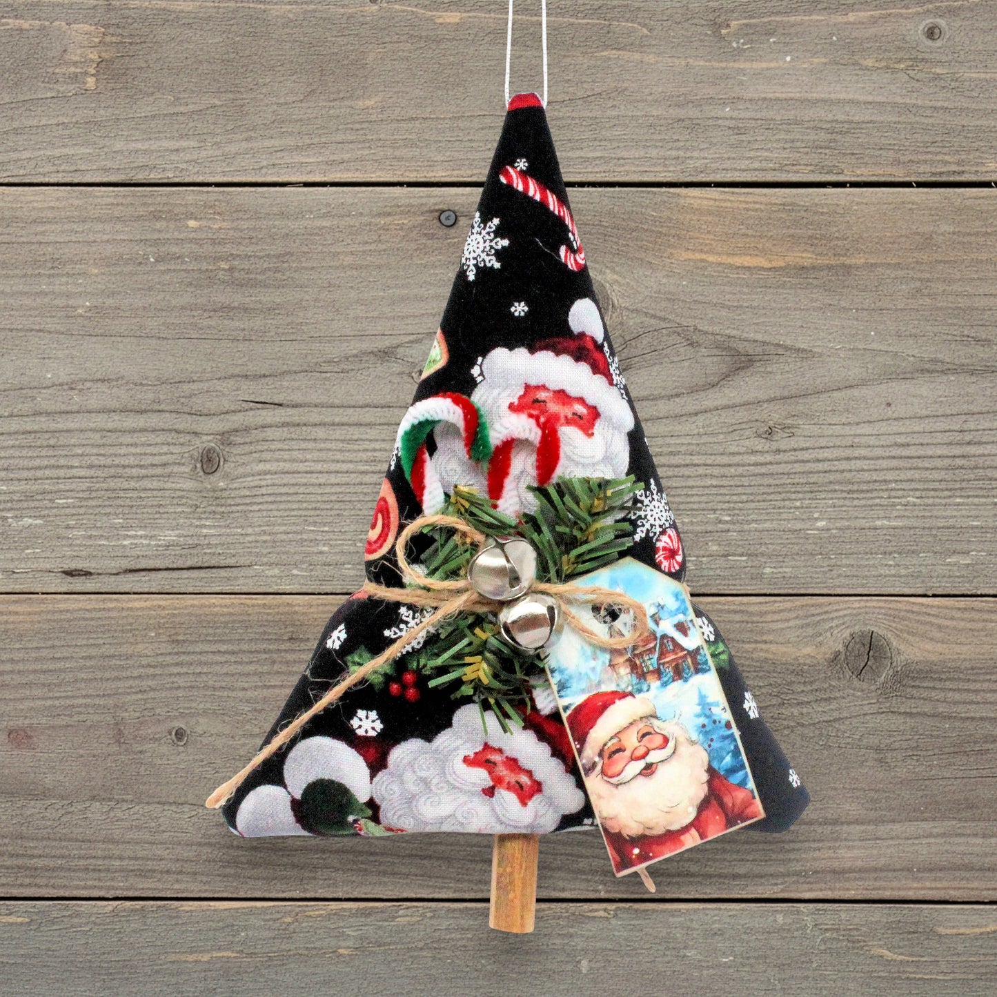 A handmade Christmas tree ornament made from black fabric with a Santa Claus and snowflake pattern, decorated with green pine branches, small silver bells, a jute twine bow, and a Santa card, hanging against a rustic wooden plank background.