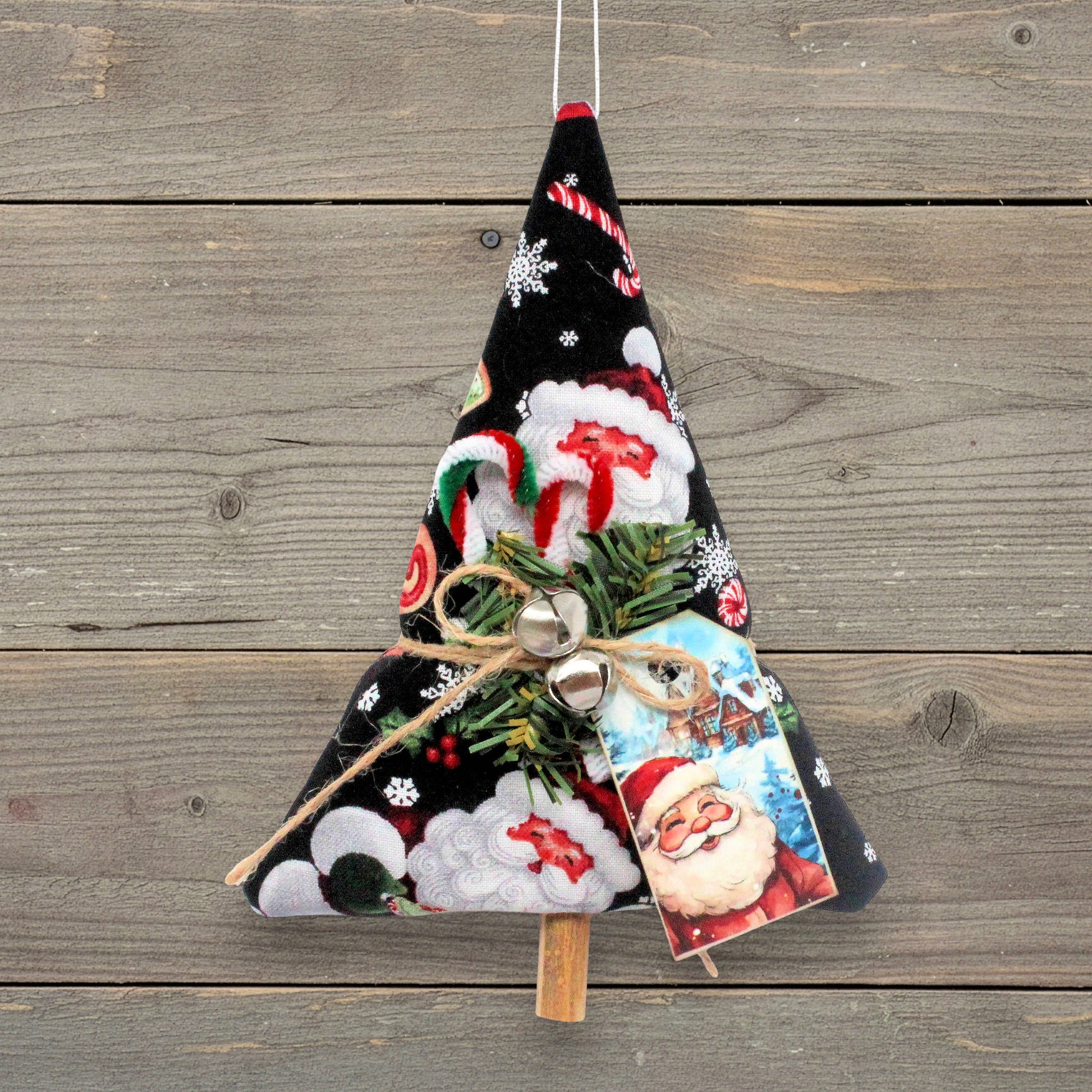 A handmade Christmas tree ornament made from black fabric with a Santa Claus and snowflake pattern, decorated with green pine branches, small silver bells, a jute twine bow, and a Santa card, hanging against a rustic wooden plank background.