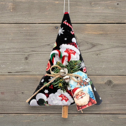 A handmade Christmas tree ornament made from black fabric with a Santa Claus and snowflake pattern, decorated with green pine branches, small silver bells, a jute twine bow, and a Santa card, hanging against a rustic wooden plank background.