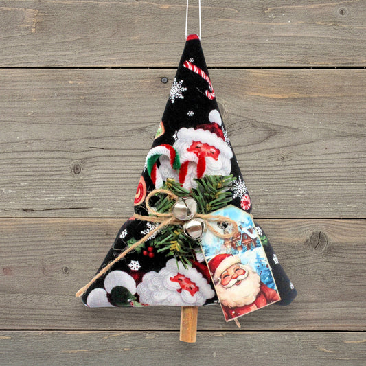A handmade Christmas tree ornament made from black fabric with a Santa Claus and snowflake pattern, decorated with green pine branches, small silver bells, a jute twine bow, and a Santa card, hanging against a rustic wooden plank background.
