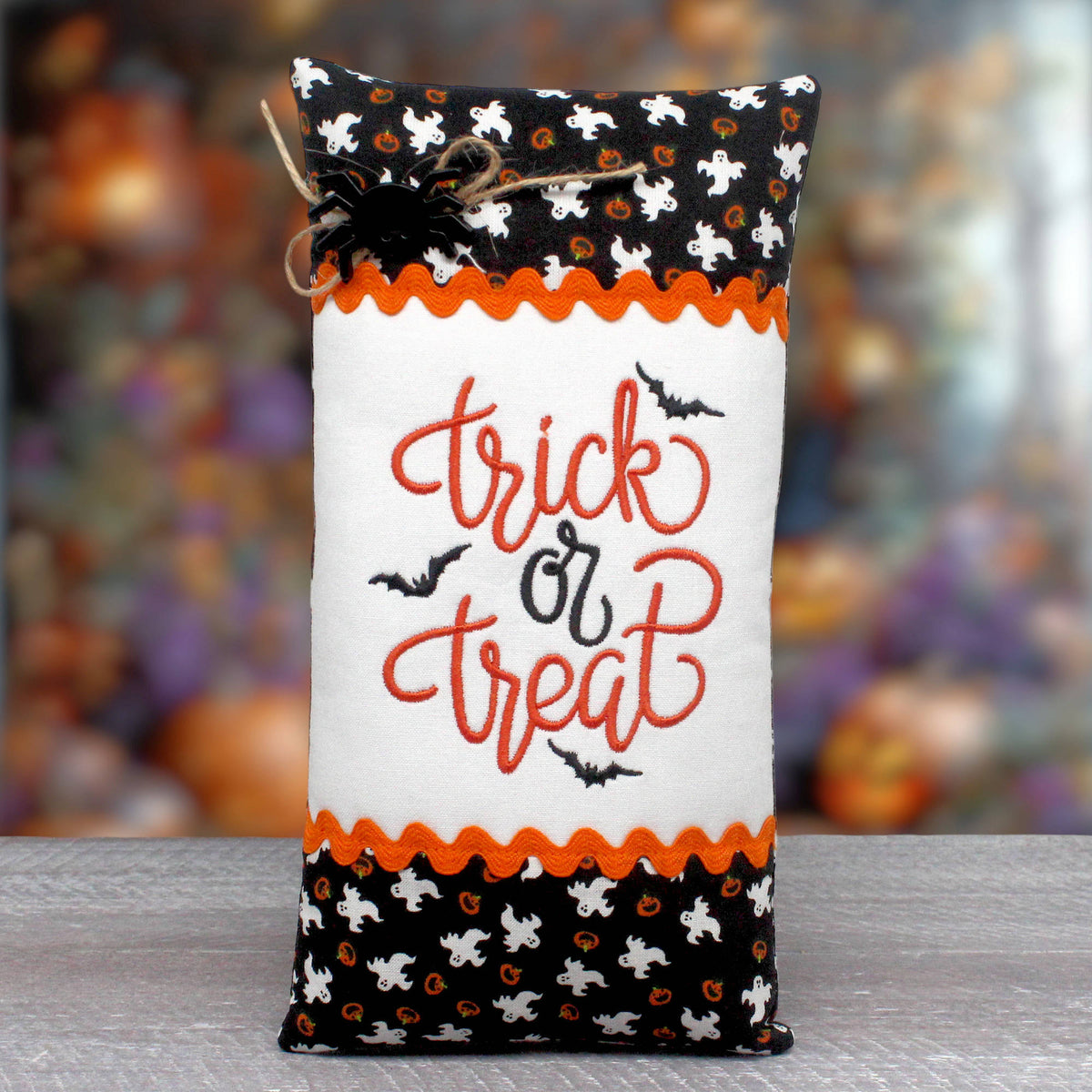 A Halloween-themed fabric pillow with 'trick or treat' embroidered in orange and black on a white center panel, with black and white patterned borders and small bat silhouettes. The bag is photographed standing upright on a gray surface against a blurred autumnal background.