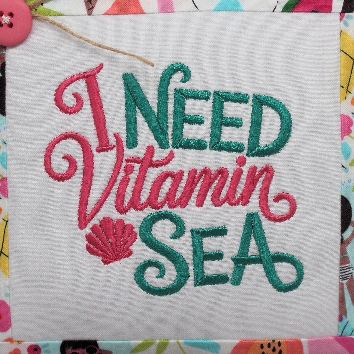 Embroidered fabric wall art with the phrase “I NEED Vitamin SEA” in pink and teal thread on a white square canvas, shown with a colorful patterned border and a pink button at the top left.