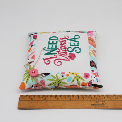 A small square fabric pillow with a colorful tropical print border and a white center panel that reads 'I Need Vitamin SEA' in stylized lettering. A pink button and twine tie are attached on the left side.
