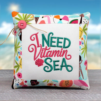 A square decorative throw pillow with a colorful tropical and beach-themed border and a central white panel embroidered with the phrase 'I NEED Vitamin SEA' in pink and teal lettering. The pillow is shown on a gray wooden surface with a blurred beach background.