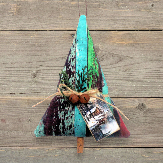 A handmade fabric ornament or made from distressed turquoise, green, black, and purple wood-painted panels, tied with twine and rusty bells, with a small printed photo tag hanging from it, set against a rustic wooden plank background.