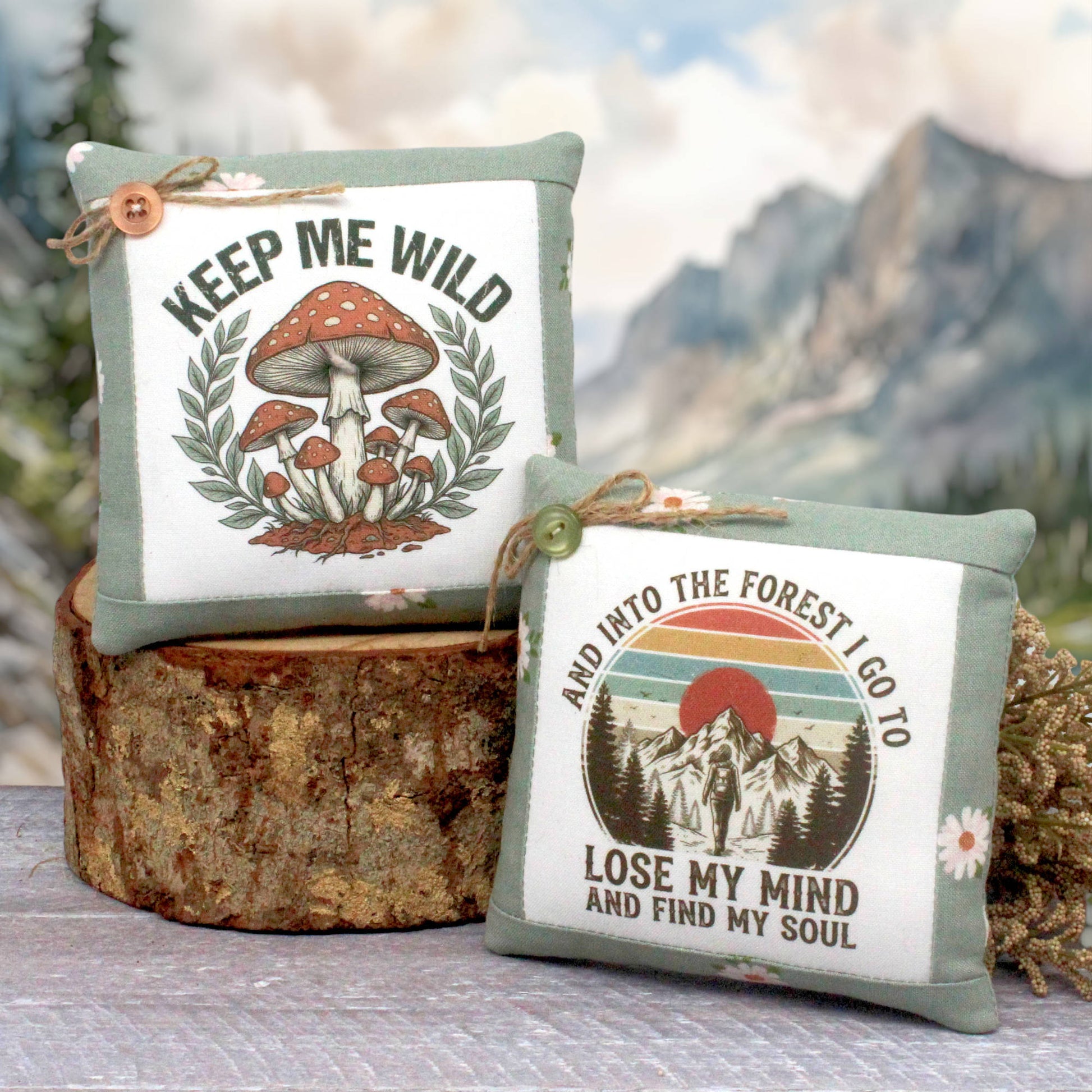 Two decorative square mini pillows displayed on a rustic wood slice with a blurred mountain forest background. The left pillow has a sage green border, a white center, a mushroom illustration, and the text 'KEEP ME WILD' with a small wooden button.