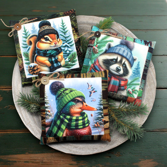 Three decorative mini pillows featuring animals wearing winter hats and scarves on a wooden surface.